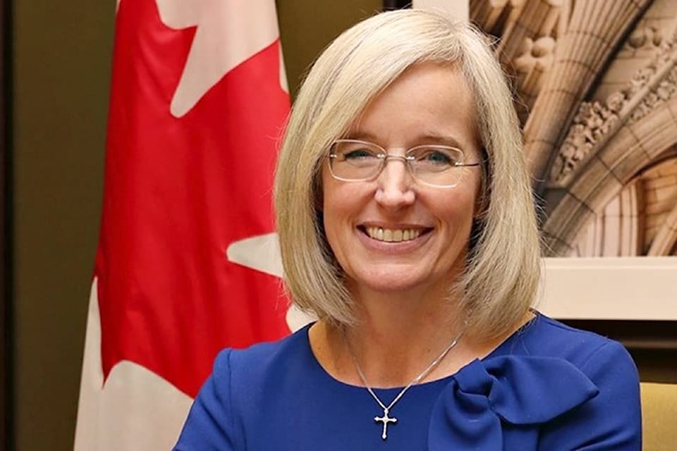 A photo of a blonde woman, Tamara Jansen, wearing glasses and a cross necklace in front of a Canadian flag