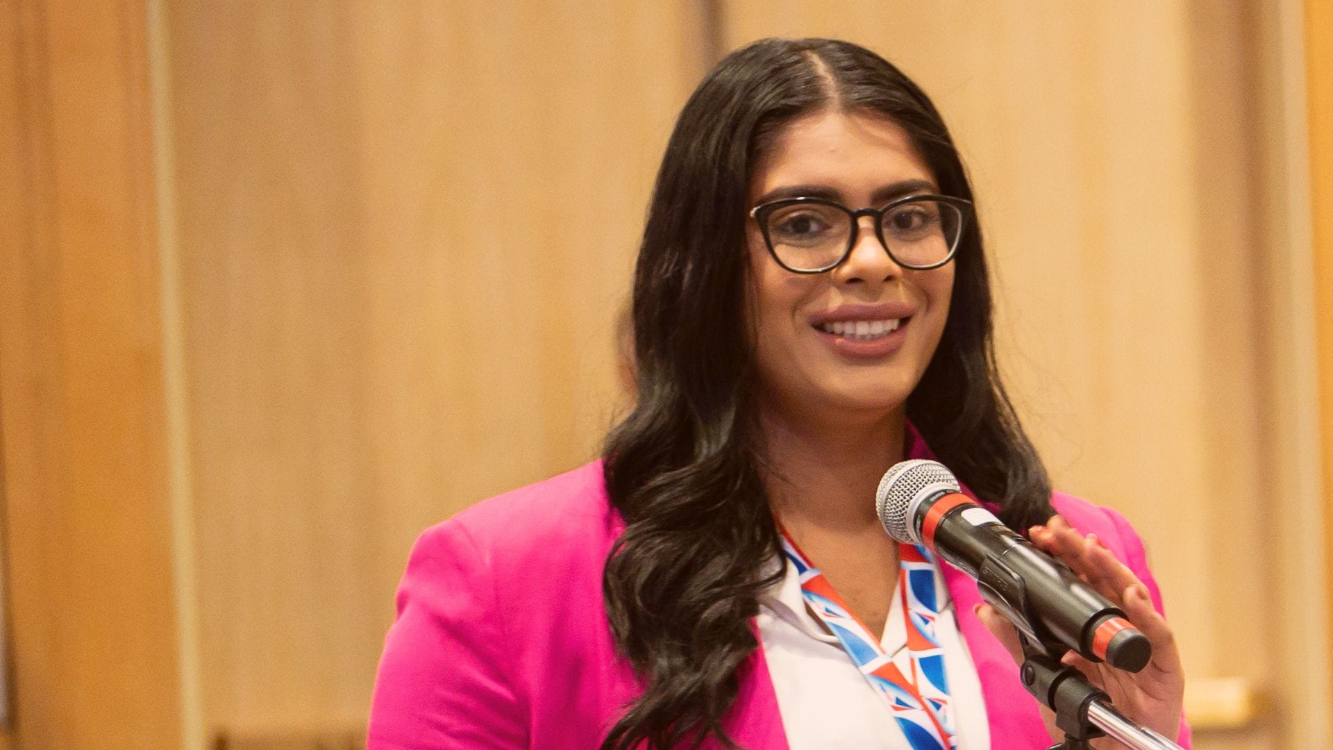 A photo of a woman, Vanessa Sharma, wearing glasses and a pink jacket in front of a microphone